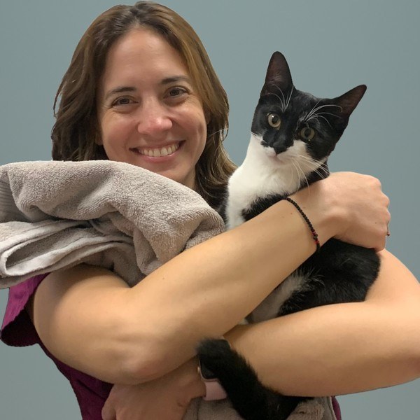 A smiling woman with shoulder-length brown hair in maroon scrubs hugs a black and white cat and a gray towel against a teal wall. A smiling woman with shoulder-length brown hair in maroon scrubs hugs a black and white cat and a gray towel against a teal wall.