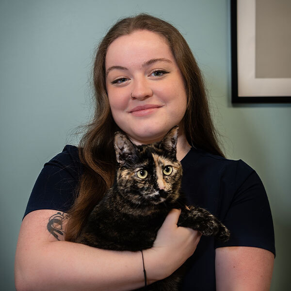 A woman with long brown hair smiles while holding a tortoiseshell cat in her arms, standing against a light blue wall with part of a framed picture visible in the background. A woman with long brown hair smiles while holding a tortoiseshell cat in her arms, standing against a light blue wall with part of a framed picture visible in the background.