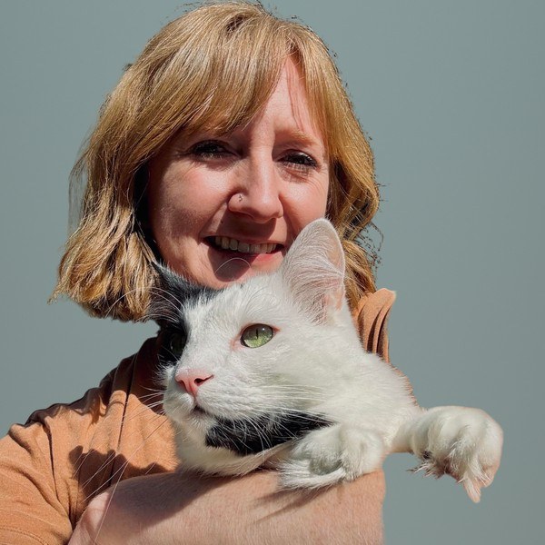 A smiling woman with strawberry-blonde hair and bangs in a rust-colored shirt holds a fluffy white cat with gray markings against a teal wall. A smiling woman with strawberry-blonde hair and bangs in a rust-colored shirt holds a fluffy white cat with gray markings against a teal wall.